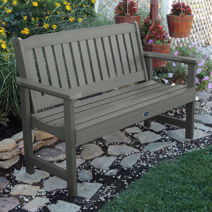 5ft light gray Blue Ridge garden bench on stone pathway with flowers in background 