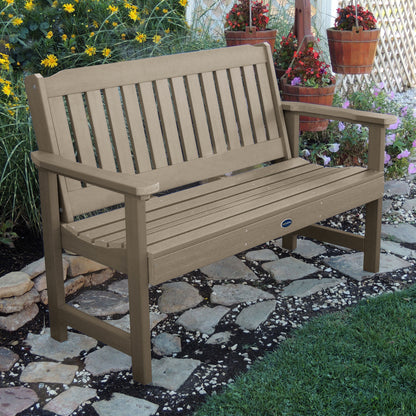 5ft light brown Blue Ridge garden bench on stone pathway with flowers in background 